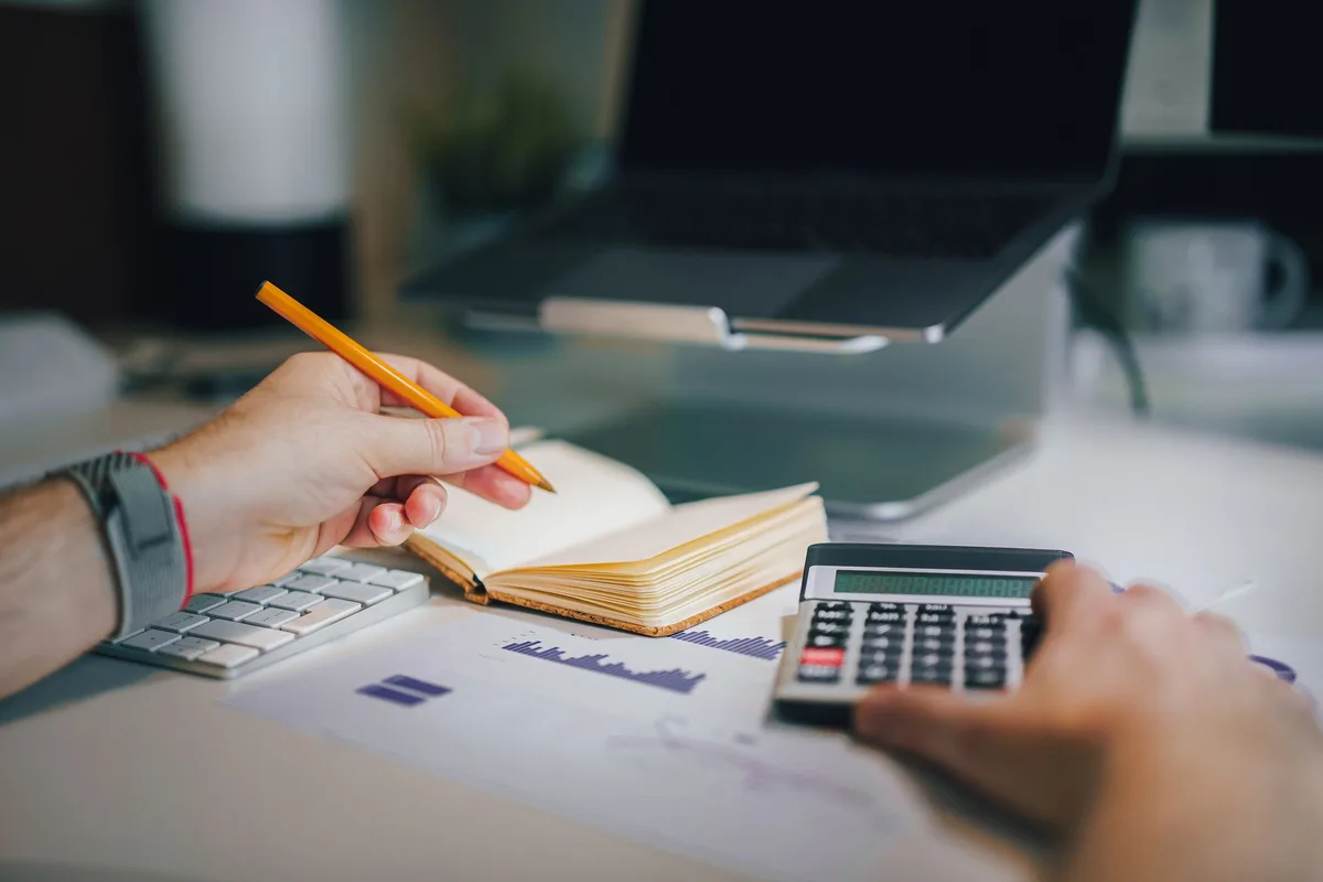 Hands using a calculator and taking notes on a desk beside a laptop while working out take-home pay